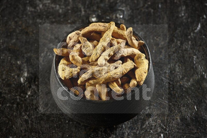 A black pottery bowl full of dried turmeric whole fingers on textured black flooring