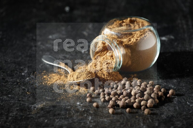 A flipped glass spice jar and metal spoon full of allspice powder and powder spilled out of it with whole balls on a textured black flooring