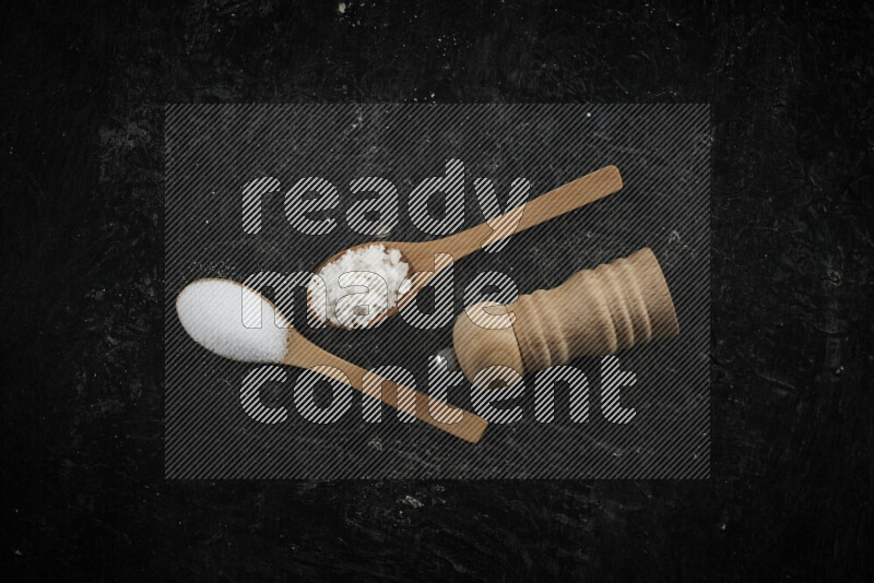 2 wooden spoons filled with fine and coarse salt with wooden grinder beside them on black background