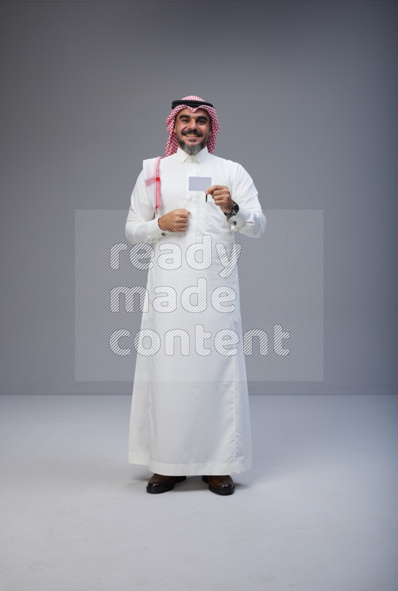 Saudi man Wearing Thob and red Shomag standing holding ATM card on Gray background