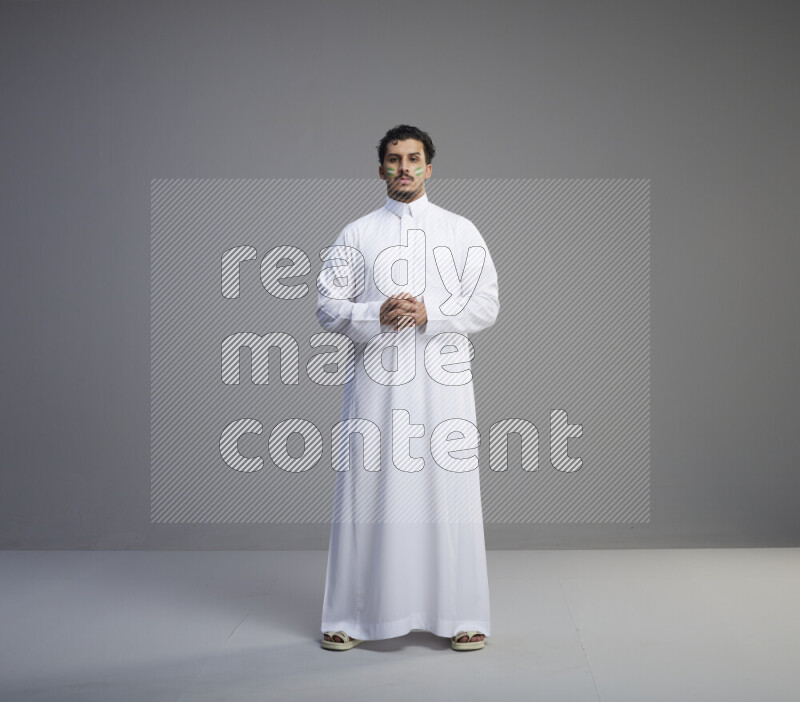A Saudi man standing wearing thob with face painting interacting with the camera on gray background