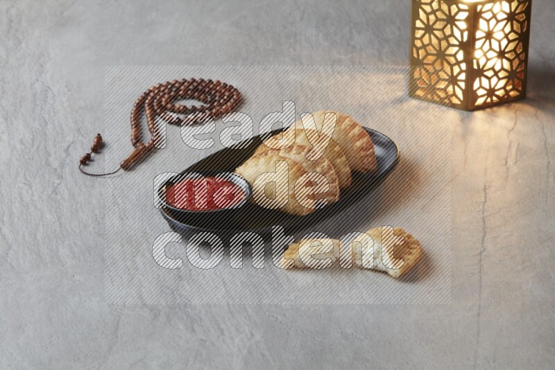 Four fried sambosas in an oval shaped black plate, beside a cut cheese sambosa, a brown misbaha and a golden lantern on a gray background