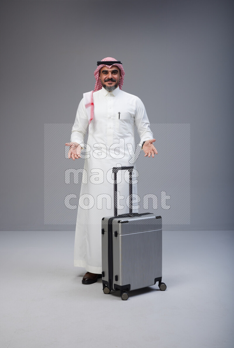 Saudi man wearing Thob and red Shomag standing holding Travel bag on Gray background