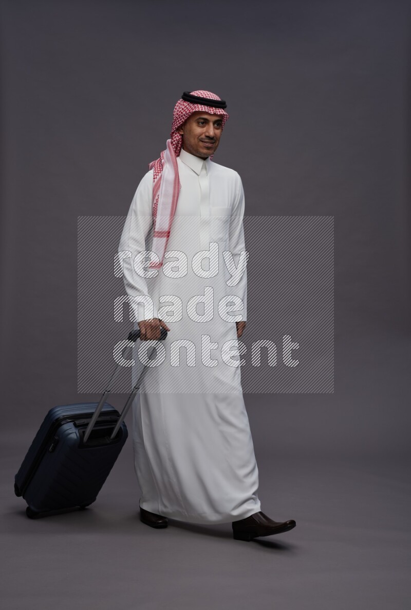 Saudi man wearing thob and shomag standing holding bag on gray background