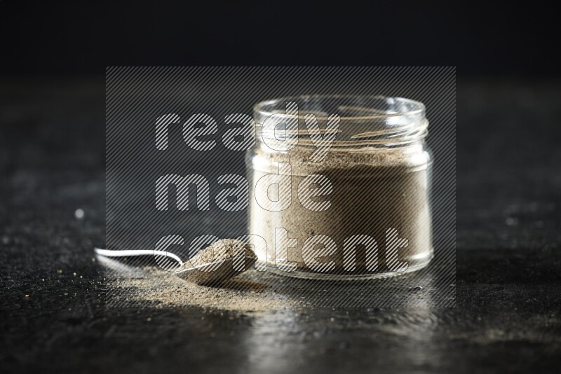 A glass jar and metal spoon full of cardamom powder on textured black flooring