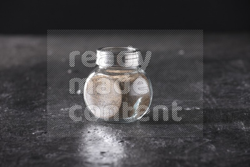 A glass spice jar full of black pepper powder on textured black flooring