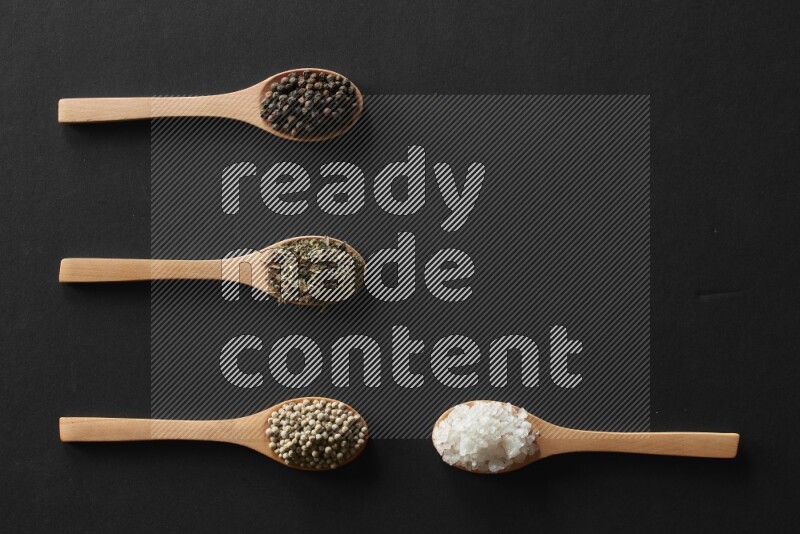 wooden spoons filled with white peppers, cloves, cardamom, salt, black peppers and basil on black flooring