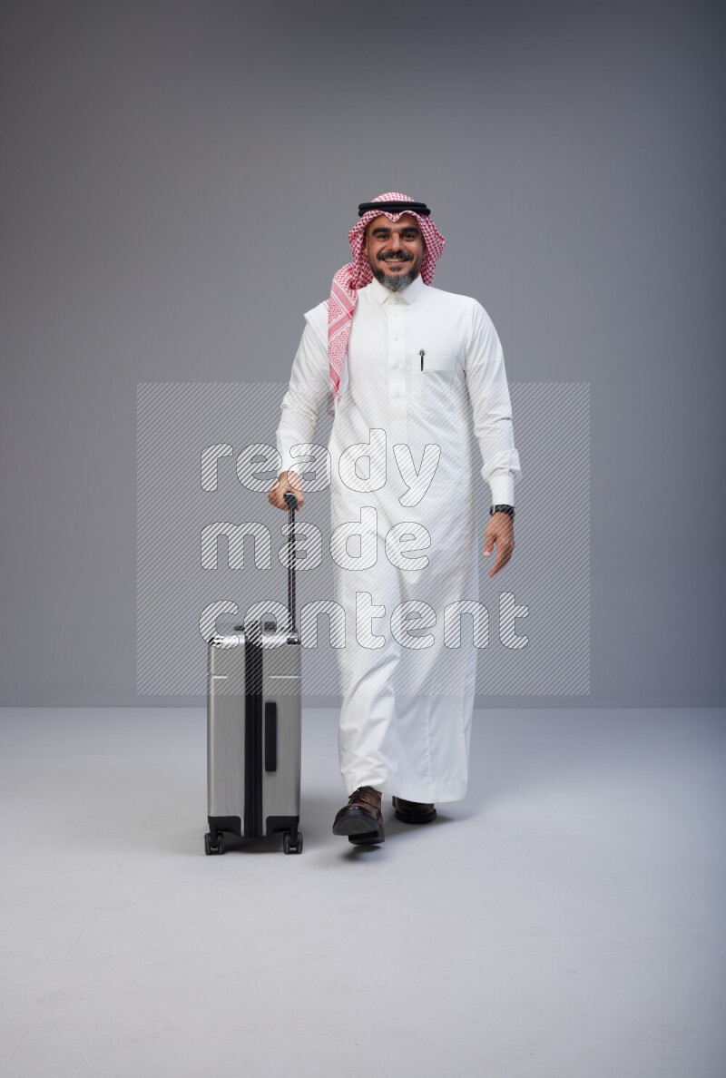 Saudi man wearing Thob and red Shomag standing holding Travel bag on Gray background