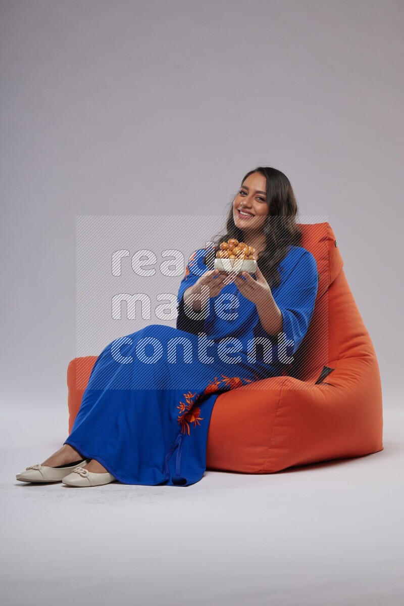 A Woman sitting on an orange beanbag wearing Jalabeya holding a plate of luqaimat