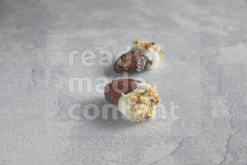 Two pecan stuffed date covered with white chocolate and chopped walnuts on alight grey background