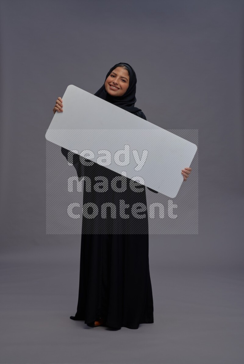 Saudi woman wearing Abaya standing holding white board on gray background