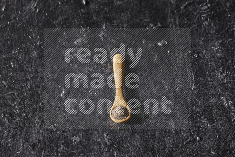 A wooden spoon full of black pepper powder on a textured black flooring