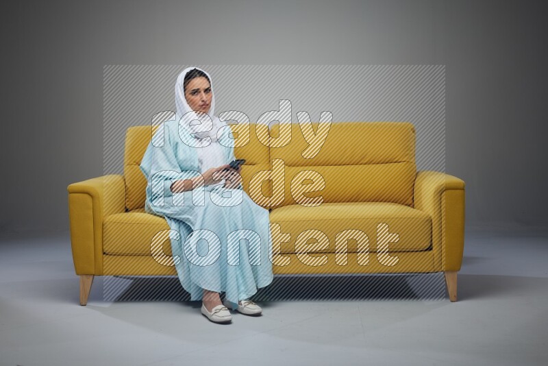 A Saudi woman wearing a light blue Abaya and white head scarf sitting on a yellow sofa and using her phone eye level on a grey background