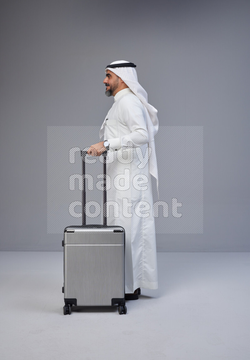 Saudi man wearing Thob and white Shomag standing holding Travel bag on Gray background