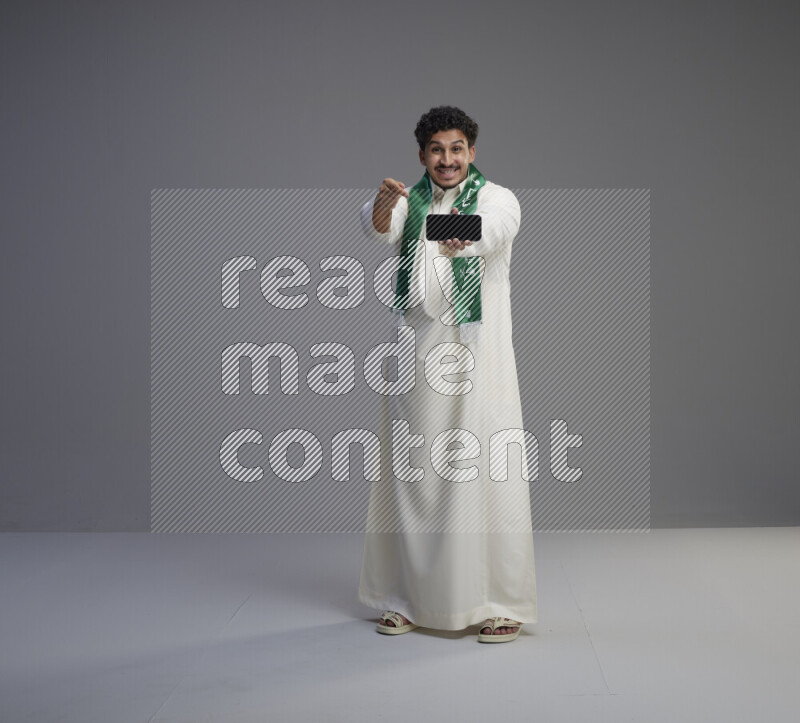 A Saudi man standing wearing thob and Saudi flag scarf showing phone to camera on gray background
