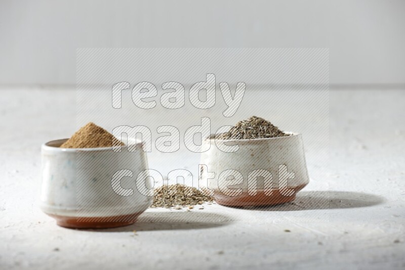 2 beige bowls full of cumin seeds and powder with spilled powder and seeds on textured white flooring