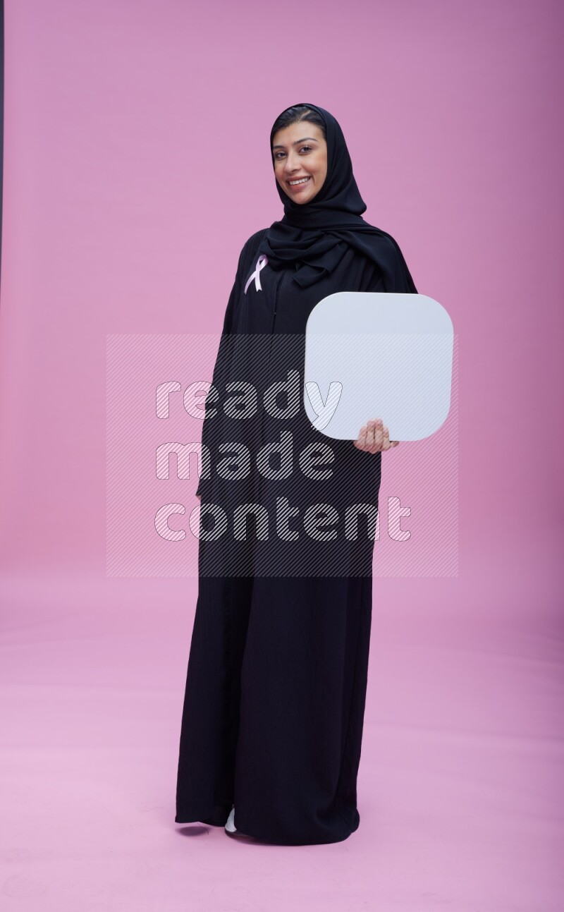 Saudi woman wearing pink ribbon on Abaya standing holding social media signs on pink background