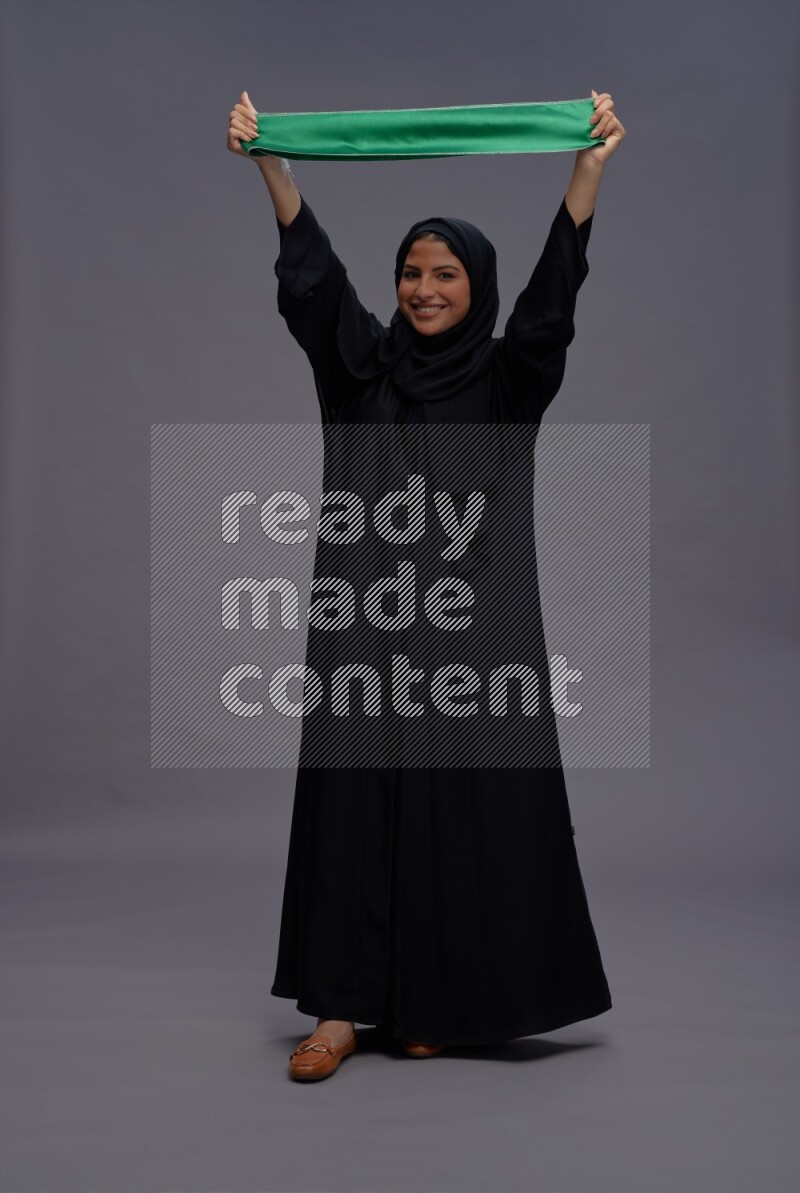 Saudi woman wearing Abaya standing holding Saudi flag on gray background