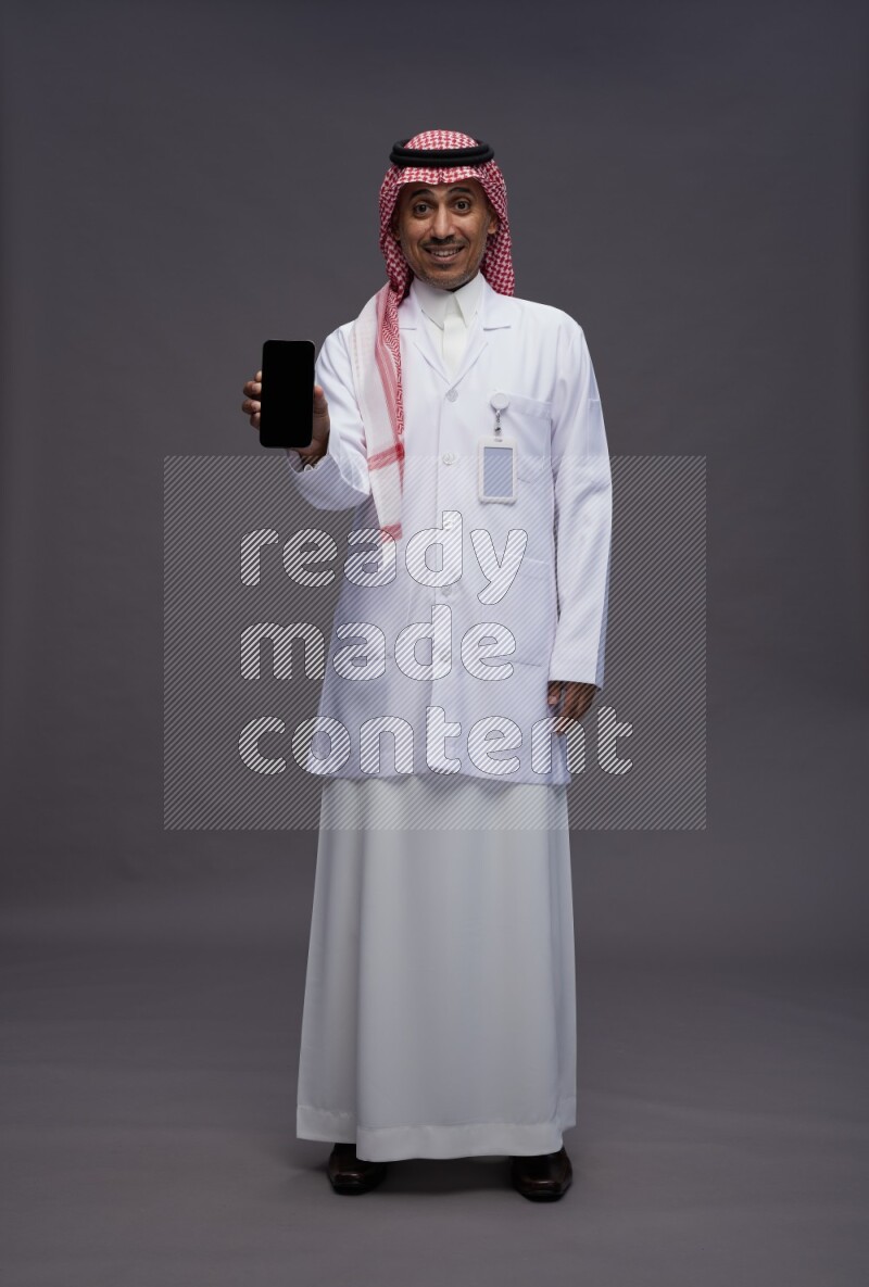 Saudi man wearing thob with lab coat and shomag with pocket employee badge standing showing phone to camera on gray background