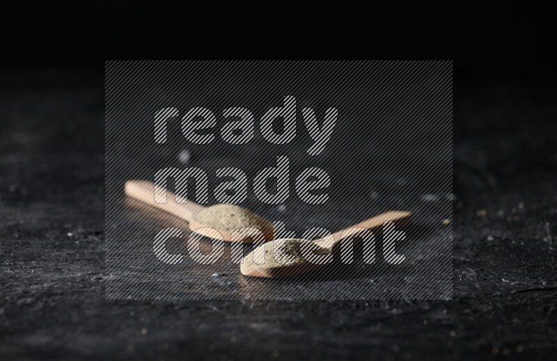 2 wooden spoons full of white pepper powder on textured black flooring