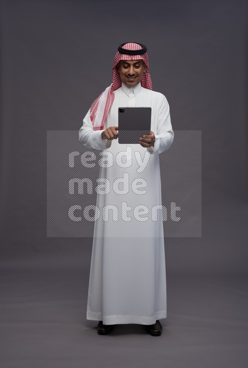 Saudi man wearing thob and shomag standing working on tablet on gray background