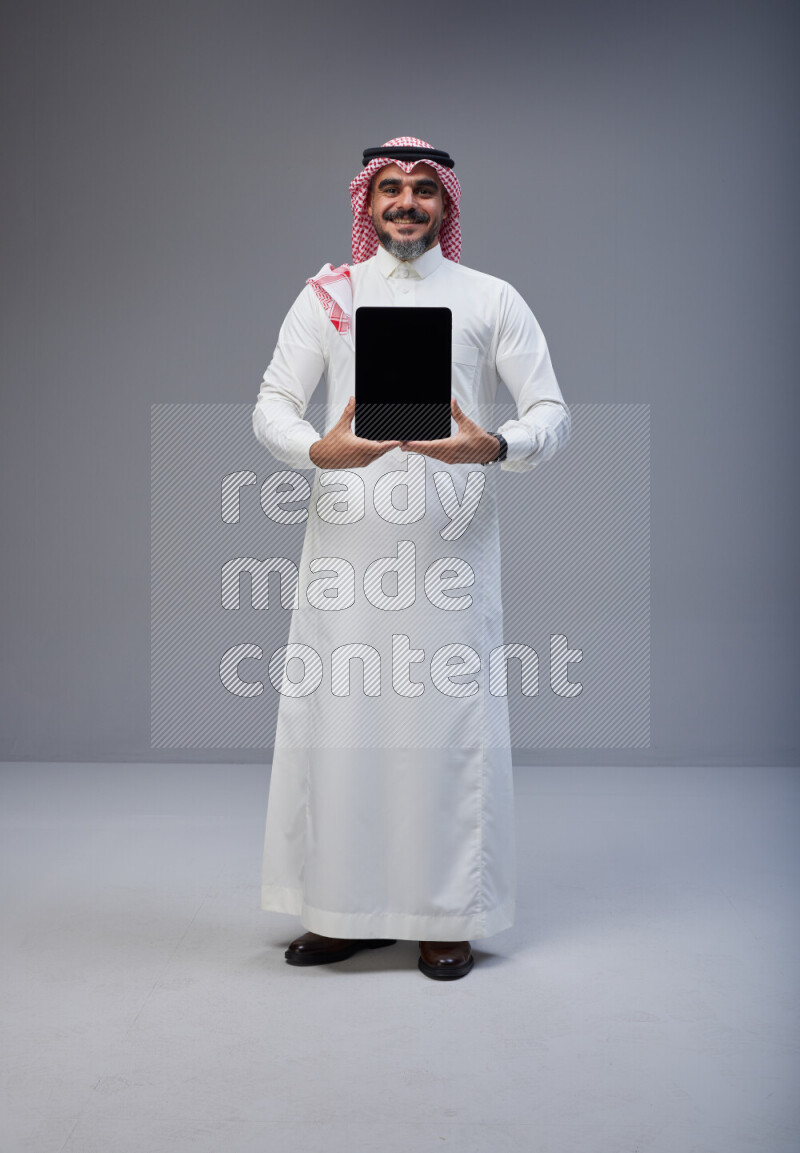 Saudi man Wearing Thob and red Shomag standing showing tablet to camera on Gray background