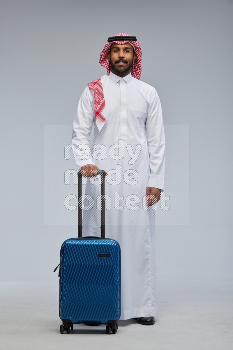 Saudi man wearing Thob and shomag standing holding Travel bag on Gray background