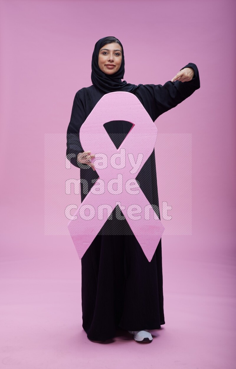 Saudi woman wearing pink ribbon on abaya standing holding awareness ribbon on plain pink background