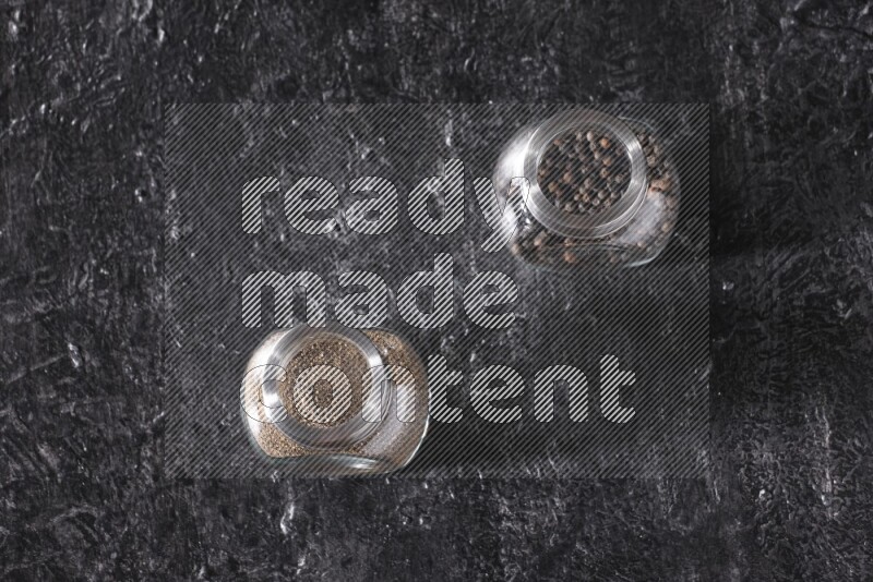 2 glass spice jars full of black pepper powder and black pepper beads on textured black flooring