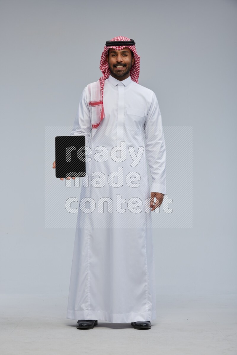 Saudi man Wearing Thob and shomag standing showing tablet to camera on Gray background