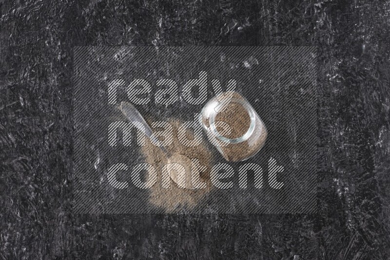 A glass spice jar full of black pepper powder and a metal spoon full of powder on textured black flooring