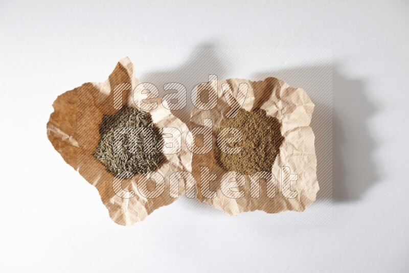 Cumin seeds and cumin powder in 2 crumpled pieces of paper on white flooring