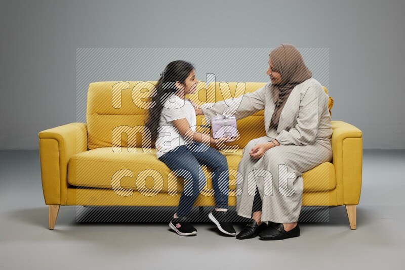 A girl sitting on a yellow sofa and giving a gift to her mother on gray background