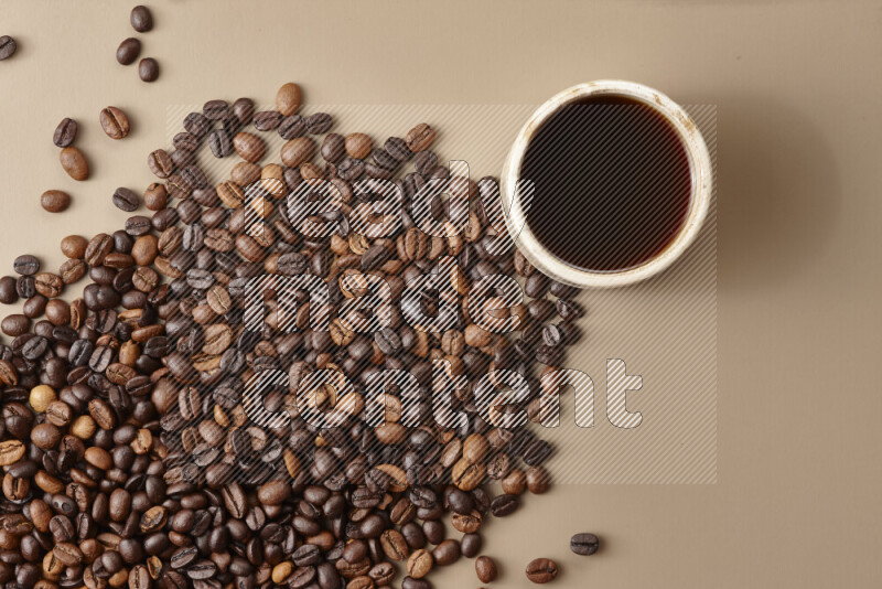 A beige pottery cup of coffee surrounded by roasted coffee beans on beige background