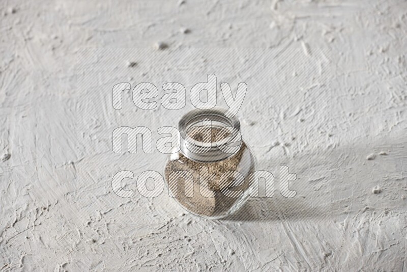 A glass spice jar full of powder black pepper on textured white flooring