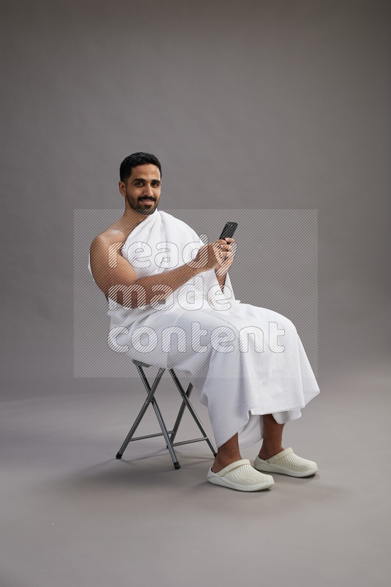 A man wearing Ehram sitting on chair texting on phone on gray background