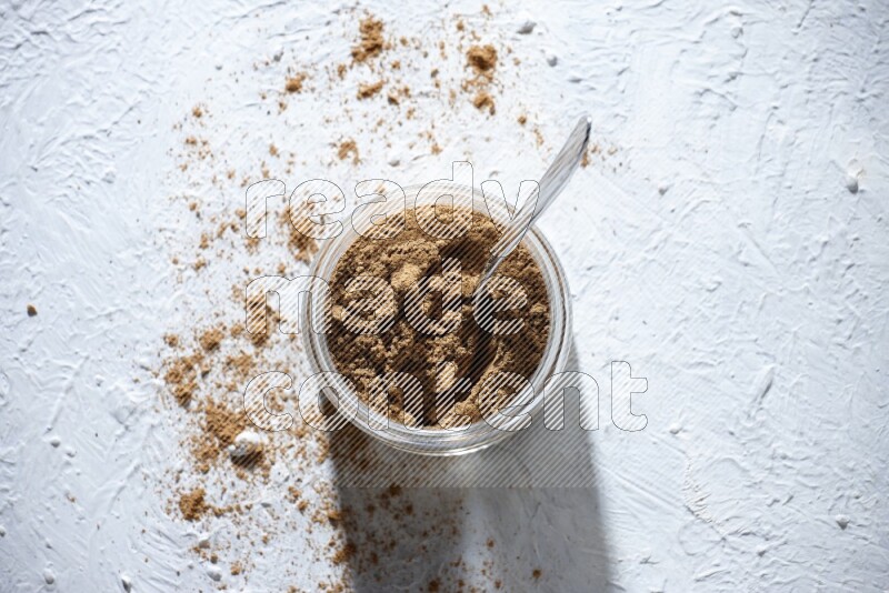 A glass jar and a metal spoon full of allspice powder on a textured white flooring