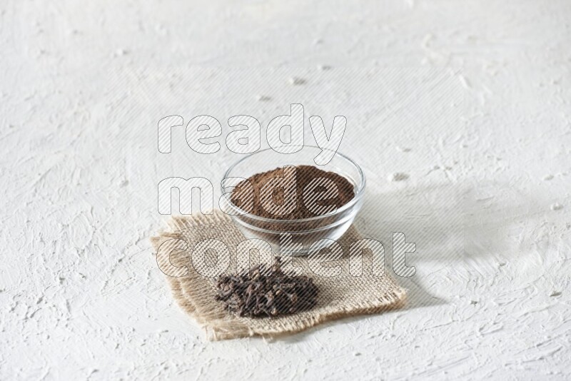 A glass bowl full of cloves powder and cloves grains on a burlap piece on a white flooring