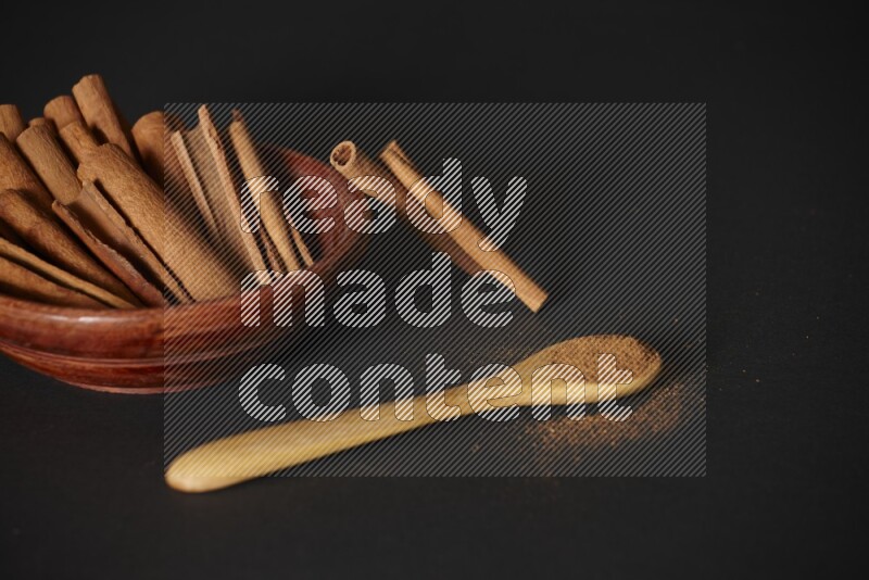 Cinnamon sticks in wooden bowl and cinnamon powder in a wooden spoon on black background