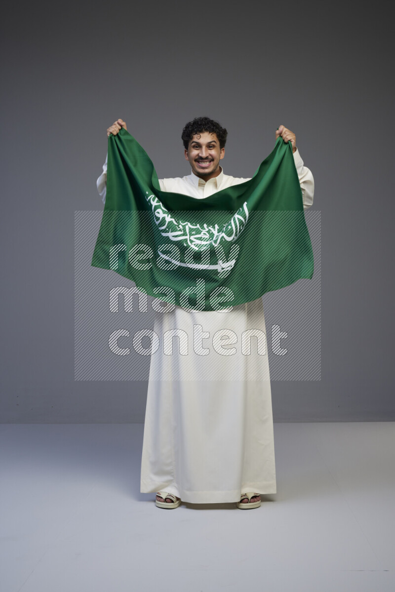 A Saudi man standing wearing thob holding big Saudi flag on gray background
