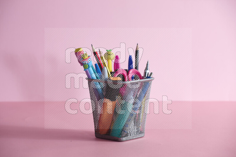 Pens, pencils and other different school supplies in a metal pen holder on rose background