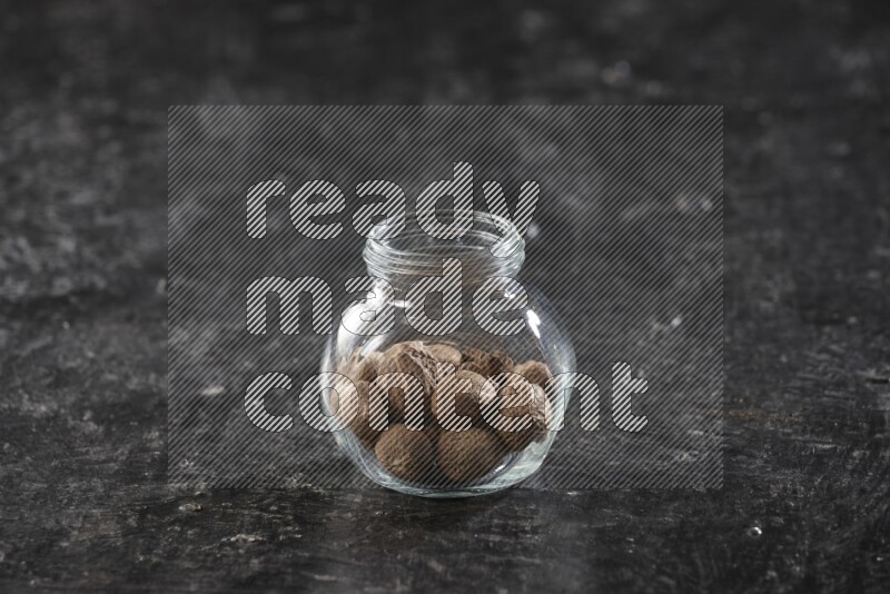 A glass spice jar full of whole nutmeg seeds on a textured black flooring