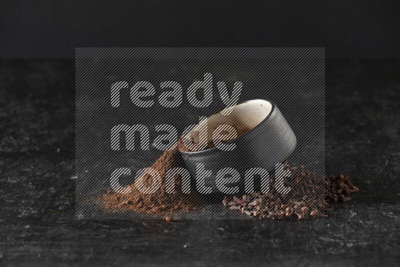 A black pottery bowl full of cloves powder spilled out of it with whole grains on a textured black flooring