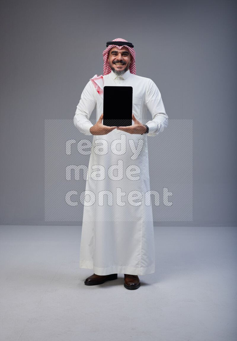 Saudi man Wearing Thob and red Shomag standing showing tablet to camera on Gray background