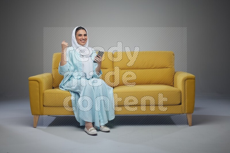 A Saudi woman wearing a light blue Abaya and white head scarf sitting on a yellow sofa and using her phone eye level on a grey background