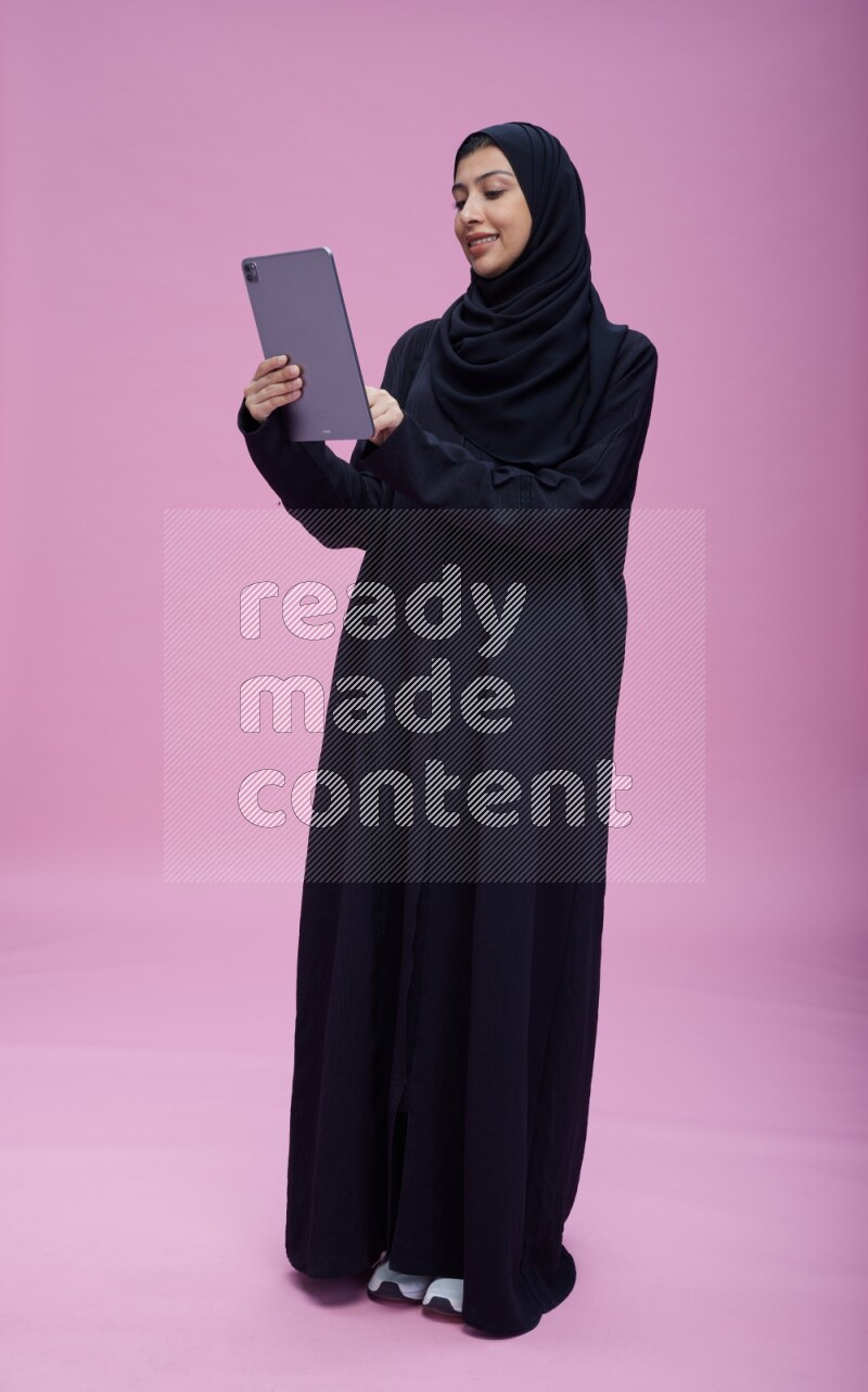 Saudi woman wearing Abaya standing working on tablet on pink background
