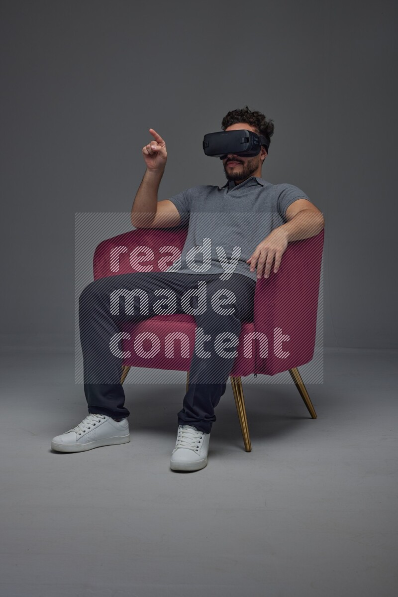 A man wearing casual and wearing VR while sitting on a burgundy chair eye level on a gray background