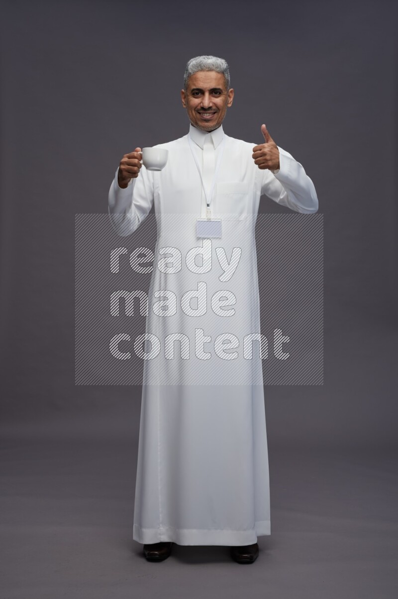 Saudi man wearing thob with neck strap employee badge standing holding mug on gray background