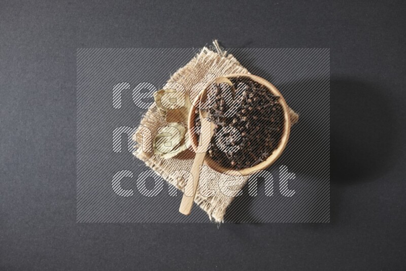 A wooden bowl, a wooden spoon full of cloves, and bay leaves (laurel) on a piece of burlap on a black flooring