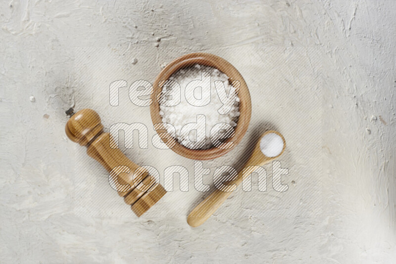 A wooden bowl and spoon filled with white sea salt and wooden grinder beside them on white background